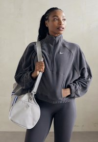 Sweatshirt cinza com fecho, de colarinho alto, apresentando um bolso na frente. Combinado com leggings pretas e uma bolsa de desporto cinza claro com detalhes em azul.