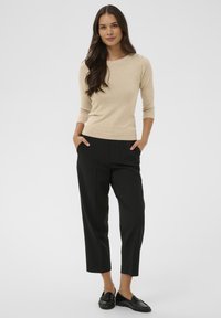 Pull beige à manches longues avec encolure ronde, associé à un pantalon noir sur mesure et des mocassins noirs, présentant une texture lisse et des lignes épurées.