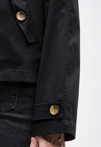 Μανίκι μαύρου τζάκετ με μεγάλο καφέ κουμπί, συνδυασμένο με σκούρο τζιν τζιν, που δείχνει ένα μέρος του χεριού σε απλό φόντο.