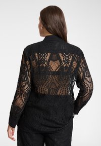 Czarna koronkowa koszula z kołnierzykiem, z misternymi wzorami i częściowo prześwitującym tyłem. Projekt uzupełniają długie rękawy i mankiety na guziki.