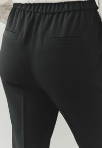 Černé zúžené kalhoty s hladkou strukturou, elastickým pasem a dvěma zadními kapsami. Design nabízí čisté linie a minimalistickou estetiku.