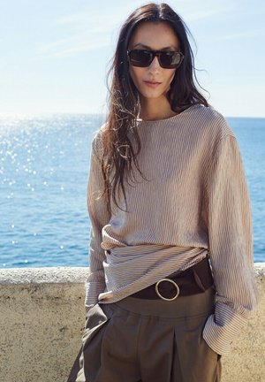 Kobieta z długimi ciemnymi włosami, nosząca ciemne okulary przeciwsłoneczne, jasną, paskowaną bluzę z długim rękawem oraz ciemne spodnie z paskiem, stojąca przy nadmurnym murze.