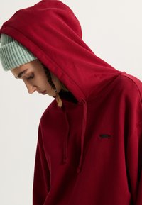 Red hoodie made of soft fabric with a large hood. Features a small black embroidered logo on the chest and a relaxed fit.
