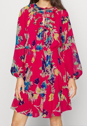 Kobieta ubrana w jasnoróżową sukienkę w kwiaty z długimi bufkami, marszczonym dekoltem i podszytą talią, sięgającą powyżej kolan.