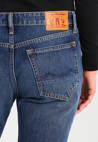 Jeans de mezclilla azul con un parche de cuero etiquetado "Kings Of Indigo". Presentan un desvanecimiento sutil, costuras en contraste y un diseño de bolsillo trasero.