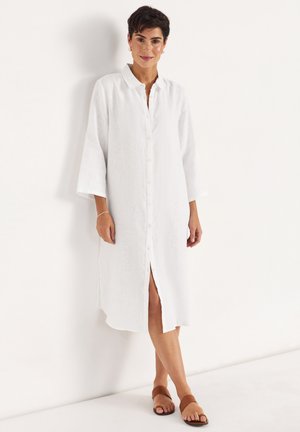 Kvinde stående op ad en væg iført en hvid langærmet skorte-kjole med knapper og brune sandaler med åbne tæer, smilende blidt.