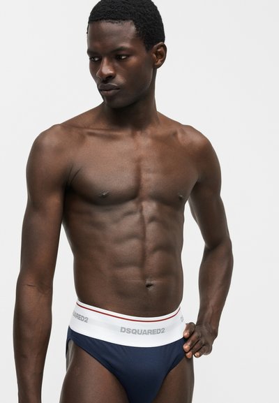 Άντρας χωρίς πουκάμισο με μυώδη κορμό, που φοράει σκούρο μπλε εσώρουχα με λευκή ζώνη DSQUARED2, κοιτάζοντας προς τα κάτω σε ένα απλό ανοιχτό φόντο.