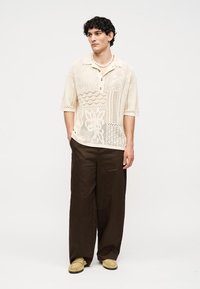 Jeune homme debout, les mains dans les poches, portant un polo en maille beige à motifs, un pantalon large marron, des mocassins beiges et un collier de perles.