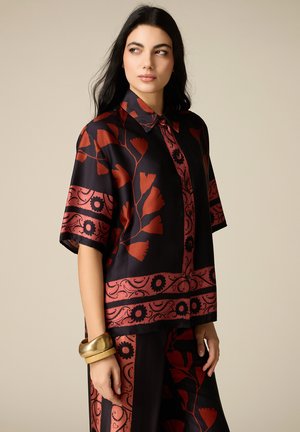 Donna che indossa una camicia a maniche corte nera e rossa con fantasia e pantaloni coordinati, con braccialetti dorati e beige al polso sinistro.