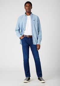 Lys blå skjorte med knapper over en hvid t-shirt, mørkeblå jeans, brun bælte og sorte lave sneakers. Glat stof og afslappet pasform.