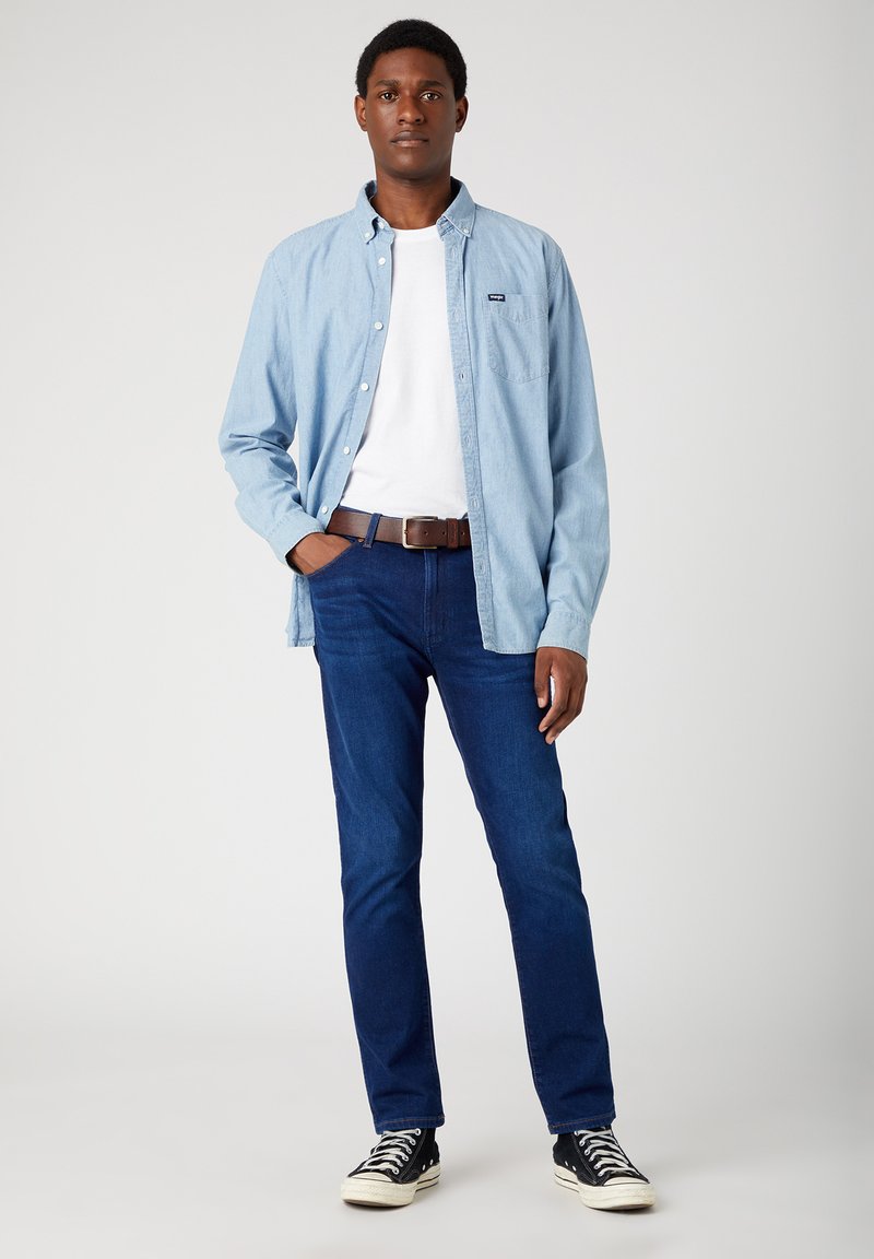 Helles blaues Hemd über einem weißen T-Shirt, dunkelblaue Jeans, brauer Gürtel und schwarze Low-Top-Sneaker. Glatte Stoffqualität und lässige Passform.