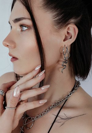 Silberne, schlangenförmige Ohrringe, geschichtete Ketten und Ringe. Lange, glänzende Nägel. Seitenprofil vor einem hellen Hintergrund. Dezentes Tattoo auf der Schulter.