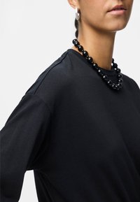 Μαύρη βαμβακερή μπλούζα με χαλαρή εφαρμογή, με κοντά μανίκια και στρογγυλή λαιμόκοψη, συνδυασμένη με μια γυαλιστερή μαύρη χάντρα.