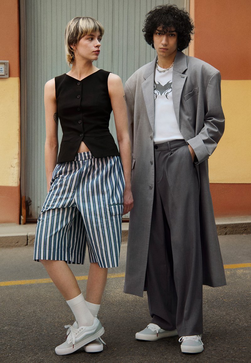 Chaleco negro recortado con botones, pantalones cortos a rayas azules y blancas, abrigo gris de gran tamaño, zapatillas blancas y calcetines grises. Un atuendo casual y moderno.
