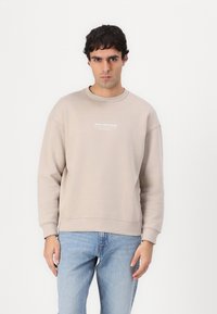 Sweatshirt bej deschis cu guler rotund, având man�șete și tiv cu nervuri. Emblema cu text alb pe piept. Purtați cu blugi albastre deschise, prezentat pe un fundal alb.