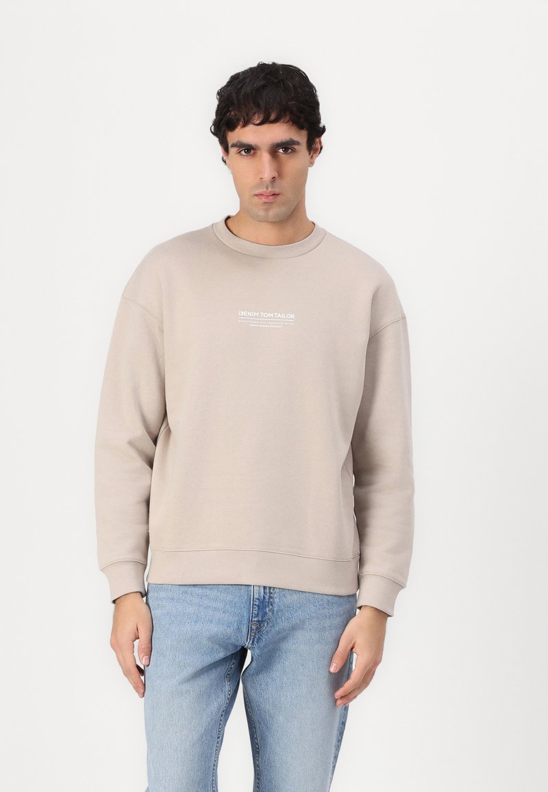 Sweatshirt bej deschis cu guler rotund, având manșete și tiv cu nervuri. Emblema cu text alb pe piept. Purtați cu blugi albastre deschise, prezentat pe un fundal alb.