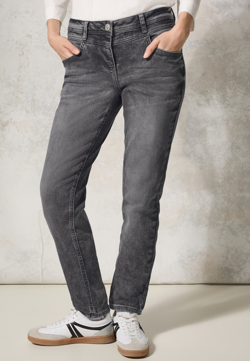 Jeans in denim grigio con vestibilità affusolata, cinque tasche, chiusura a bottone e dettagli di sbiadimento sottili. Abbinati a sneakers bianche con accenti neri.