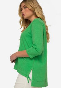 Grüner Pullover mit Dreiviertelärmeln, seitlichen Schlitzen und Bindebändern am Saum. Erhältlich in einem weichen, strukturierten Finish.