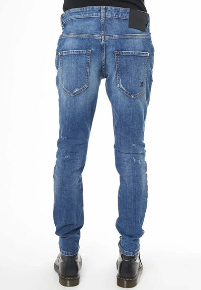 Jeans in denim blu con una vestibilità slim, caratterizzati da un'etichetta nera sulla vita, dettagli rovinati e due tasche posteriori. Stivali neri visibili.