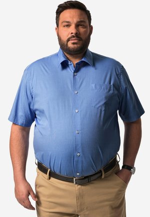 Camicia a maniche corte con colletto di colore azzurro, realizzata in tessuto morbido. Presenta una tasca frontale e chiusura con bottoni. Indossata ordinatamente dentro pantaloni khaki.