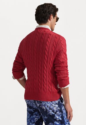 Rød kabelstrik sweater med ribbede manchetter og kant, båret over en hvid skjorte med krave. Baggrunden er ensfarvet hvid, hvilket fremhæver outfittet.