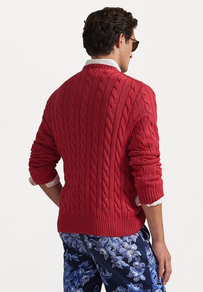 Roter kabelgestrickter Pullover mit gerippten Bündchen und Saum, getragen über einem weißen Kragenhemd. Der Hintergrund ist ganz in Weiß gehalten, was das Outfit betont.