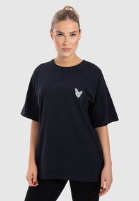 Schwarzes Baumwoll-T-Shirt mit kurzen Ärmeln und rundem Ausschnitt, das auf der linken Brustseite ein kleines graues Schmetterlingsmotiv zeigt.