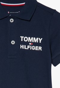 Námořnická polo košile vyrobená z texturované tkaniny. Obsahuje tříknoflíkovou légu a vyšívané logo "TOMMY HILFIGER" v bílé barvě s červeným akcentem.