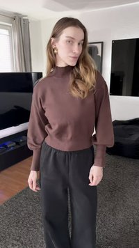 Καφέ πουλόβερ με υψηλό λαιμό, μανίκια μπαλονέ και ριμπ μανσέτες, συνδυασμένο με φαρδιά μαύρη παντελόνια. Ομαλή υφή.