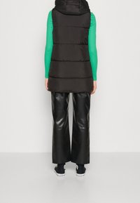 Schwarze Steppweste mit Kapuze, gestepptes Design, kombiniert mit grünem Langarmoberteil und glänzenden schwarzen weiten Hosen, getragen mit Turnschuhen.