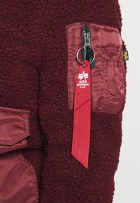 Bordová fleecová bunda s kontrastní kapsou z nylonu v barvě vínové. Má zipové zavírání a červenou stužku s logem "ALPHA INDUSTRIES U.S.A."