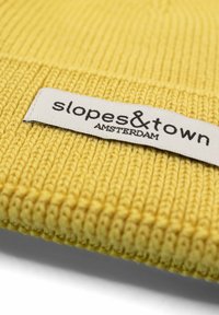 Żółta czapka beanie dzianinowa z ribbed teksturą. Posiada białą etykietę z napisem "slopes&town AMSTERDAM" wydrukowanym na czarno.