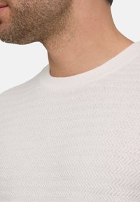 Weißer strukturierter Strickpullover mit Zickzackmuster, rundem Halsausschnitt und gerippten Bündchen. Nahaufnahme, die Material und Design betont.