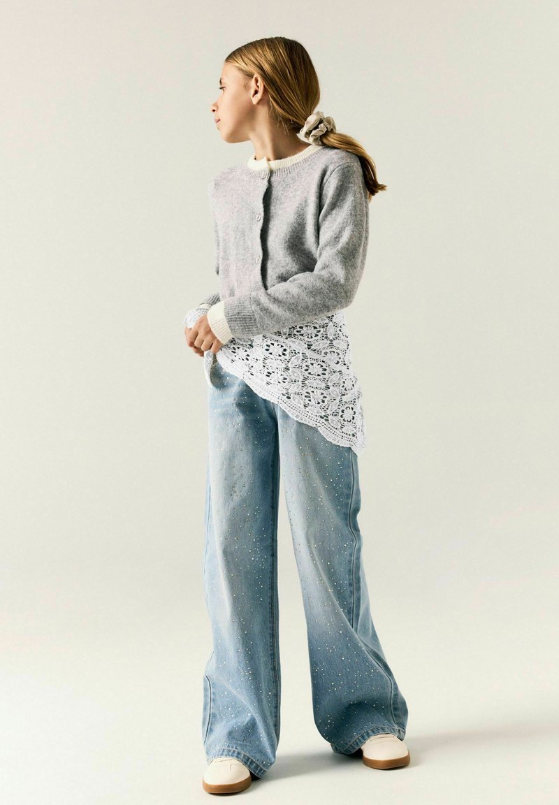 Jeune fille aux longs cheveux attachés, portant un pull gris, un haut en dentelle blanche, un jean large bleu clair avec des paillettes et des chaussures blanches, regardant vers la gauche.