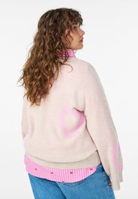 Noor naine pikkade lokkis juustega, kellel on seljas beež kampsun roosade südametega, roosade triipudega särk ja sinised teksad, küljele pööratud.