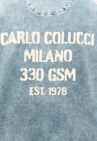 Ανοιχτό μπλε υφαντό τζιν με λευκό κείμενο που γράφει "Carlo Colucci Milano 330 GSM Est. 1978" με έντονη, φθαρμένη γραμματοσειρά.
