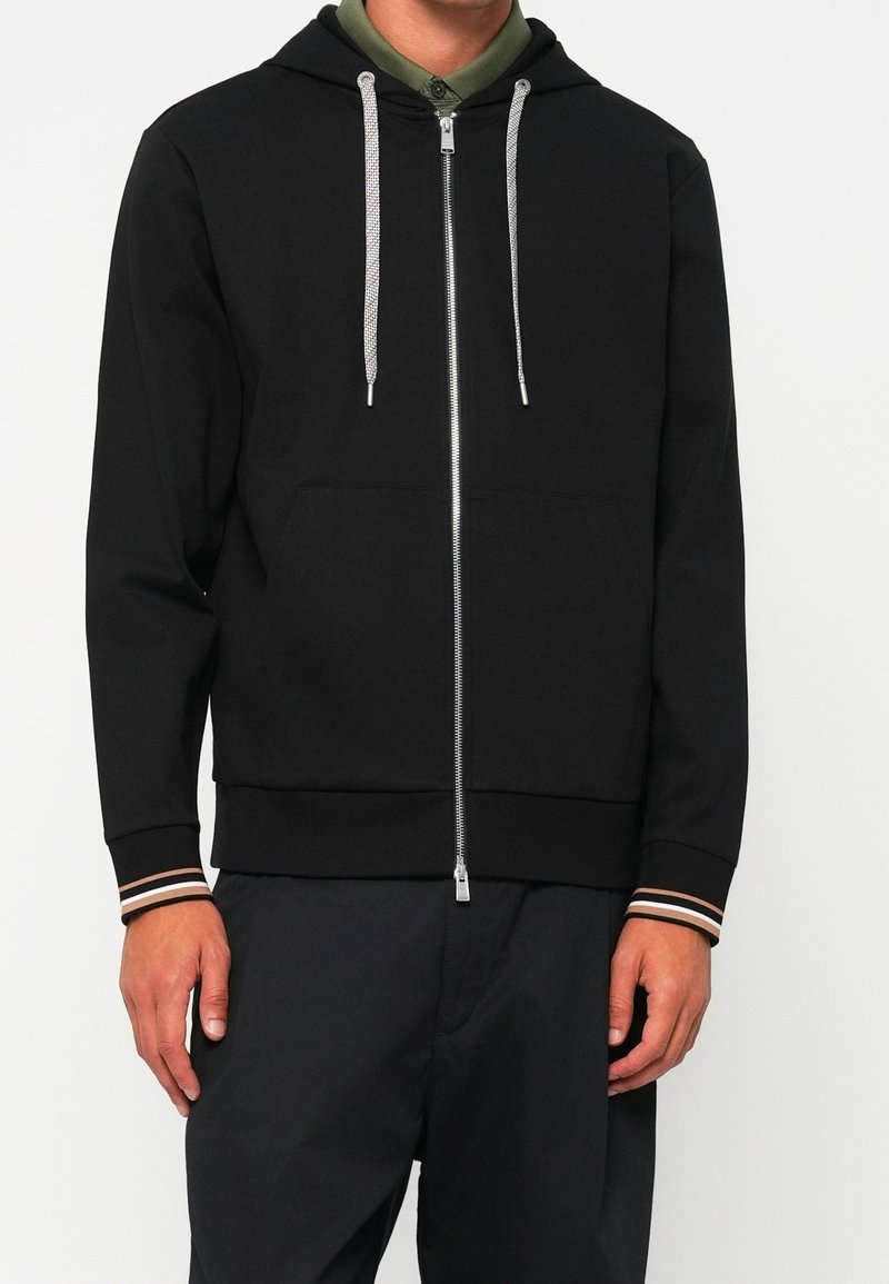 Homme portant un sweat à capuche noir zippé avec cordons blancs et poignets rayés sur une chemise verte à col, associé à un pantalon noir.