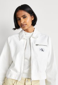 Giacca di denim bianca cropped con zip frontale, tasche laterali e un patch con il logo. Indossata sopra una blusa leggera e testurizzata e pantaloni chiari.