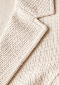 Texturovaná béžová látka s mřížkovým vzorem, s ostře vymezenými hranami a měkkým, tkaným vzhledem. Detailní záběr záhybů látky.