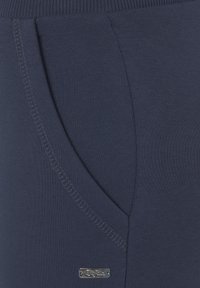 Marineblaue Jogginghose aus glattem Baumwollmaterial, mit einer Seitentasche und einem silbernen Logo-Akzent. Rippdetails am Bund.