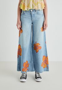 Jeans de perna larga em azul claro com padrões de flores de hibisco laranja brilhante. Combinados com ténis de lona preto e branco, proporcionando um visual casual.