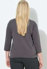 Vaaleatukkainen nainen, jolla on pitkät suorat hiukset, yllään löysä, tummanharmaa kolme neljäsosaa hihoilla varustettu paita ja istuvat tummanharmaat housut, seisoo selin katsojaan.