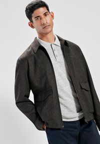 Jachetă cu fermoar de culoare maronie, cu două buzunare frontale, purtată cu un tricou polo de culoare gri deschis. Jacheta are guler și un design structurat.