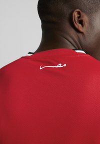 Raudonas sportinis marškinėliai, pagaminti iš kvėpuojančios medžiagos, turi juodą ir baltą apykaklę bei baltą logotipo detalę ant nugaros. Tekstas matomas ant apykaklės.