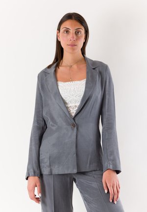 Donna che indossa un blazer grigio e pantaloni con un top in pizzo bianco, in piedi contro uno sfondo chiaro e semplice, guarda avanti con un'espressione neutra.