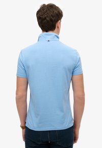 Lichtblauw poloshirt met korte mouwen en een opstaande kraag, voorzien van een klein driekleurig embleem aan de achterkant van de nek. Gladde stoftextuur.