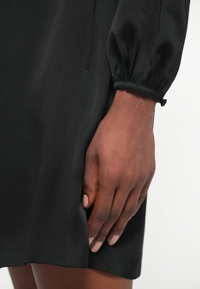 Rochie neagră cu o textură netedă, mâneci lungi și umflate, și un efect de strângere la mansete. O mână atinge tivul ținutei.