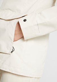 Ruka částečně uvnitř kapsy světle béžové bundy s černým patentním knoflíkem a logem "OMNI-TECH" na manžetě.