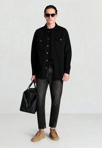 Schwarze Hemdjacke mit silbernen Knöpfen und zwei Brusttaschen, kombiniert mit dunklen Denim-Jeans und beigen Wildlederschuhen. Accessoires umfassen schwarze Sonnenbrillen und eine schwarze Tasche.