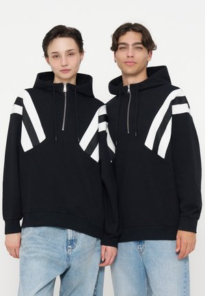 Zwei junge Erwachsene stehen nebeneinander und tragen schwarze Kapuzenpullover mit weißen diagonalen Streifen und hellblaue Jeans vor weißem Hintergrund.